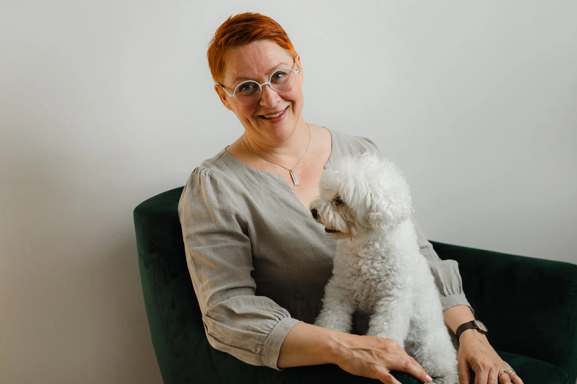 Psykoterapeutti Kati Puhakka istuu vihreässä nojatuolissa pieni valkoinen koira sylissään