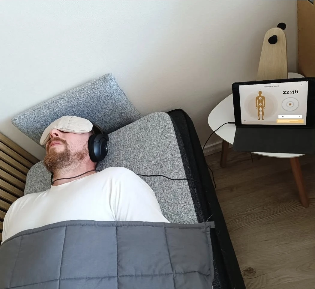 Henkilö lepää Neurosonic-patjalla kuulokkeet ja silmälaput päällä, vieressä ohjelmaa näyttävä tabletti